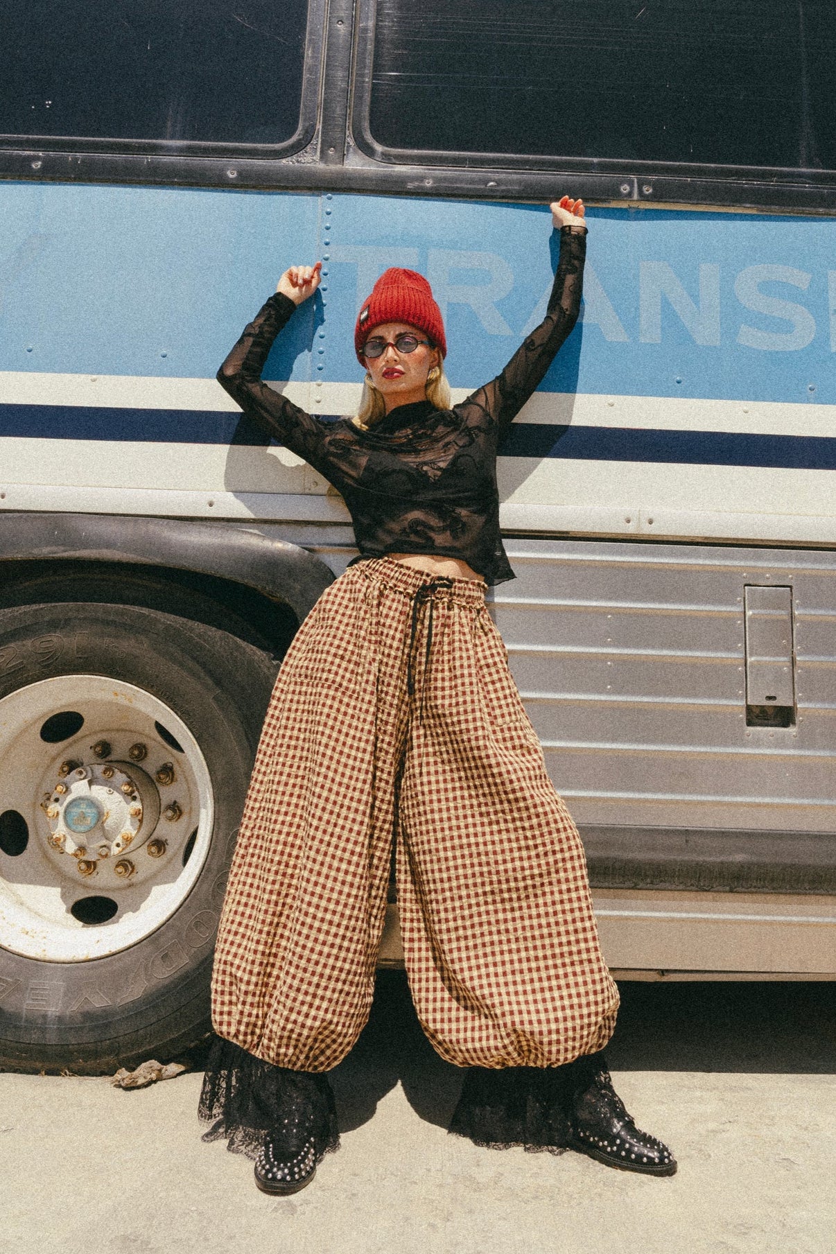 Person wearing a black top, beige checkered pants, and red beanie standing in front of a bus.