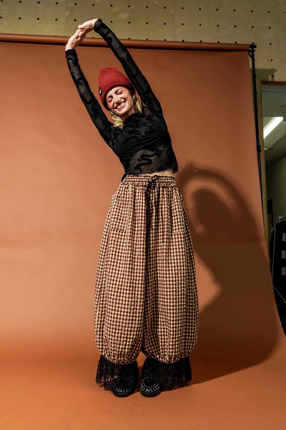 Person posing against a brown backdrop with a checkered skirt and black top.