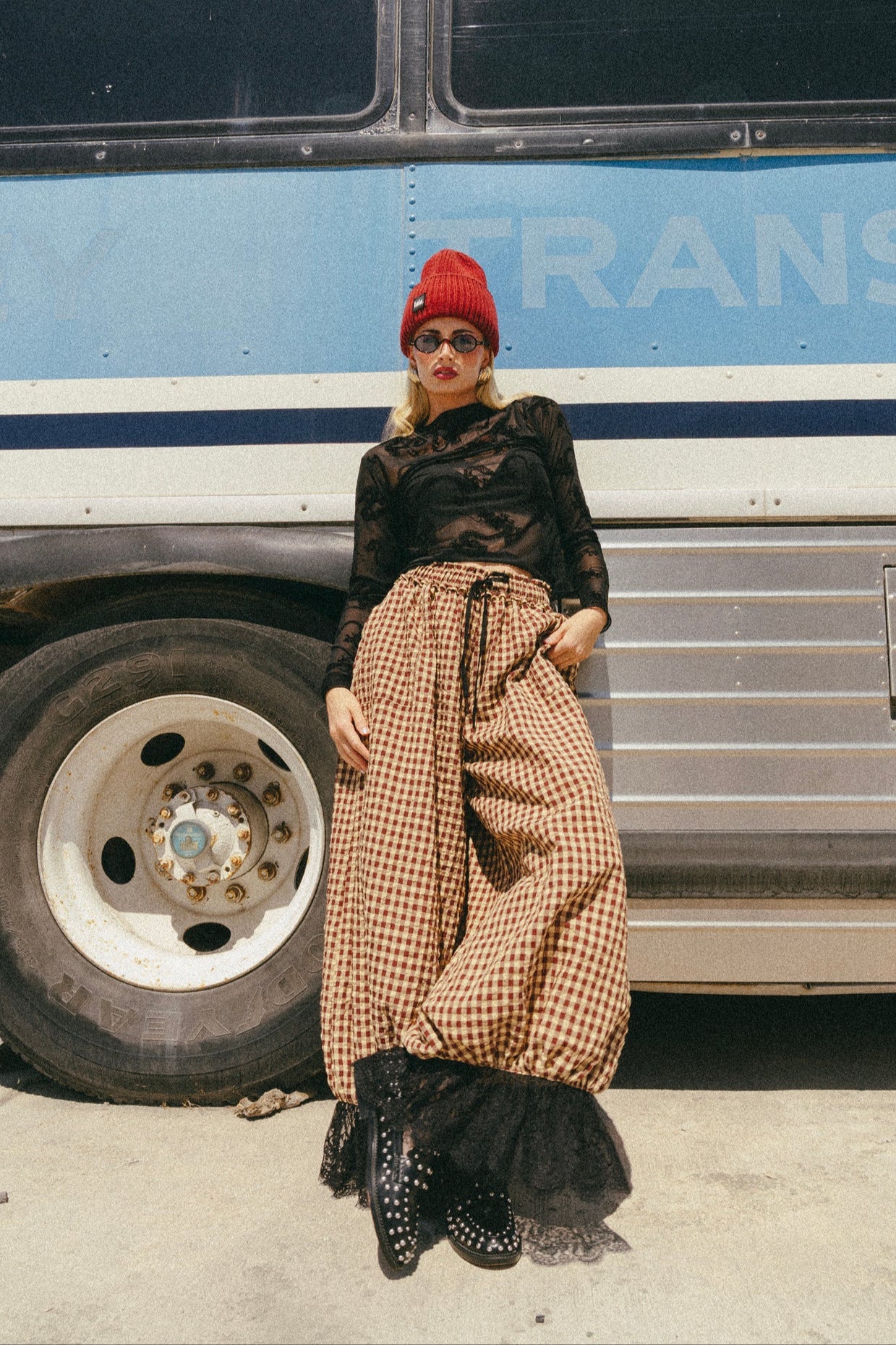 Person wearing a red beanie, black top, and checkered pants leaning against a bus with 'TRANS' on it.