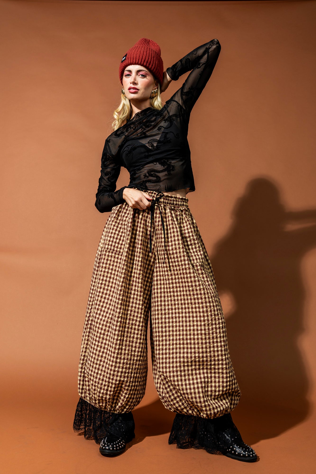 Woman wearing a black top, red beanie, and plaid pants against a brown background