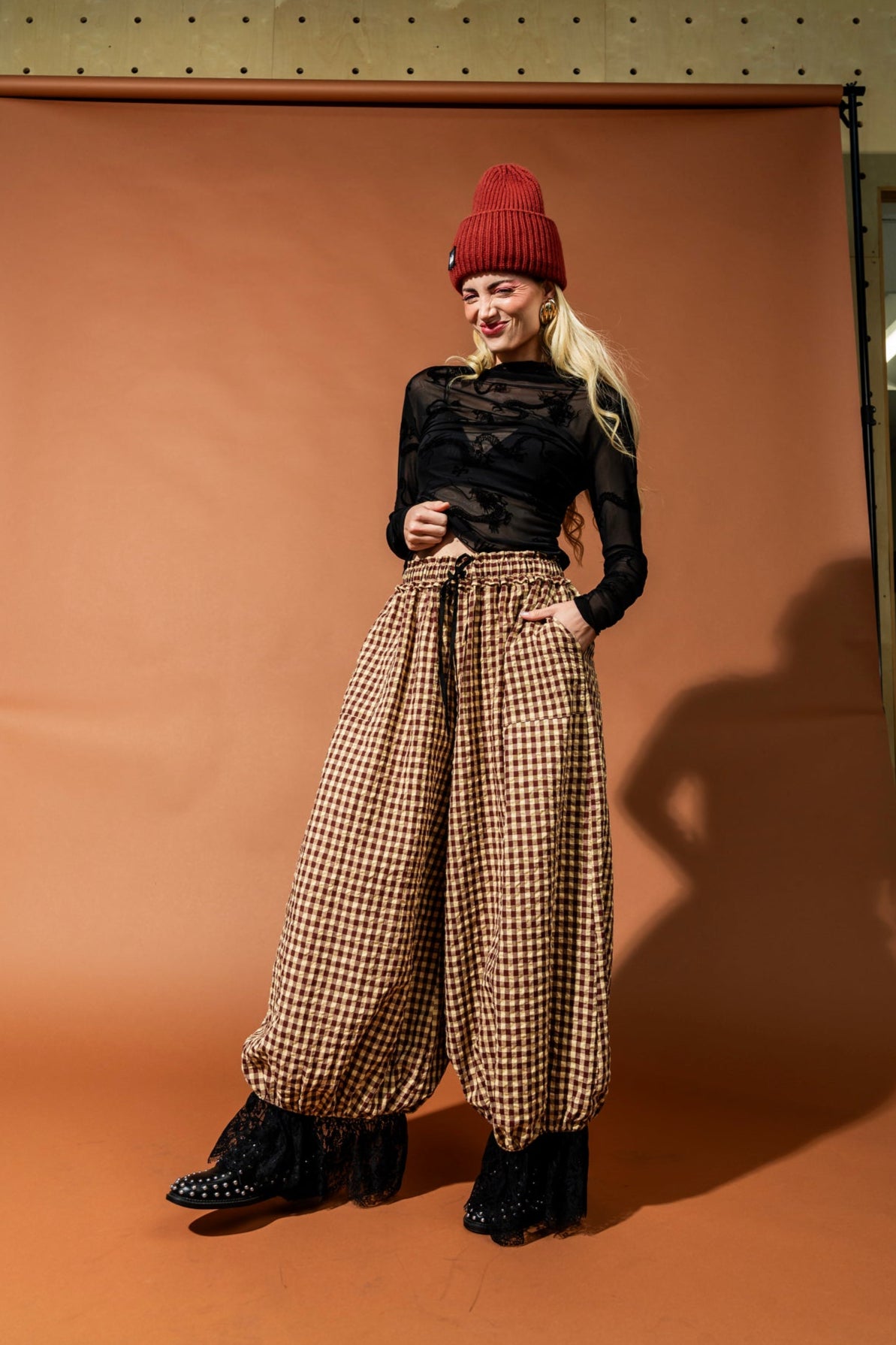 Woman wearing a black top, red beanie, and checkered pants against a brown backdrop