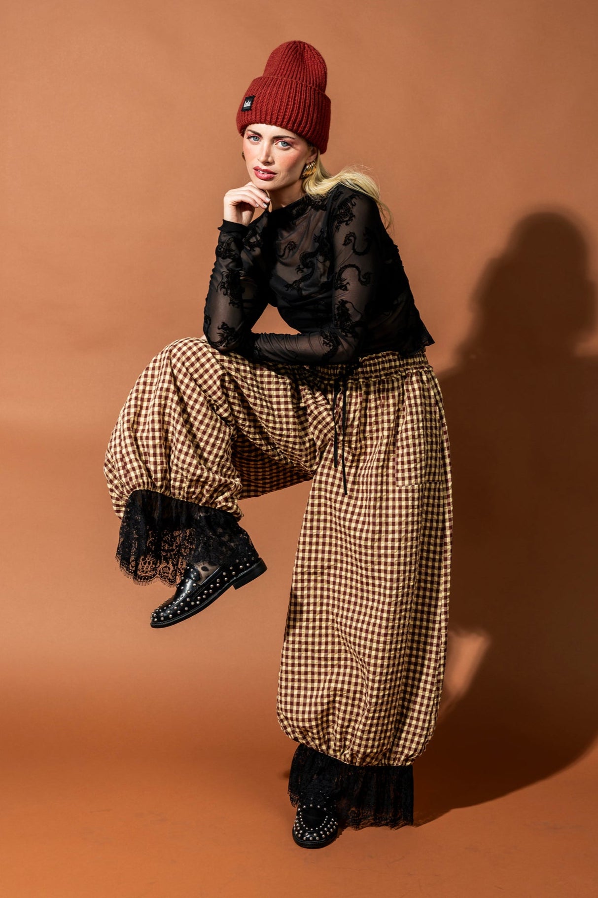 Woman wearing a black lace top and checkered pants with a red beanie on a brown background