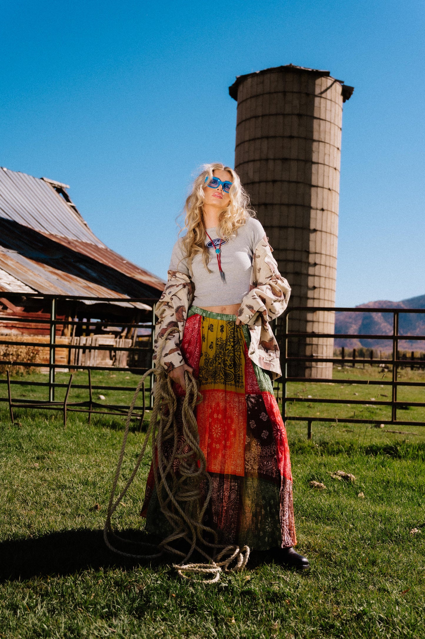 Woman standing in a rural setting with a silo and barn in the background wearing a patchwork style bandana print pleated midi skirt with an elastic waistband