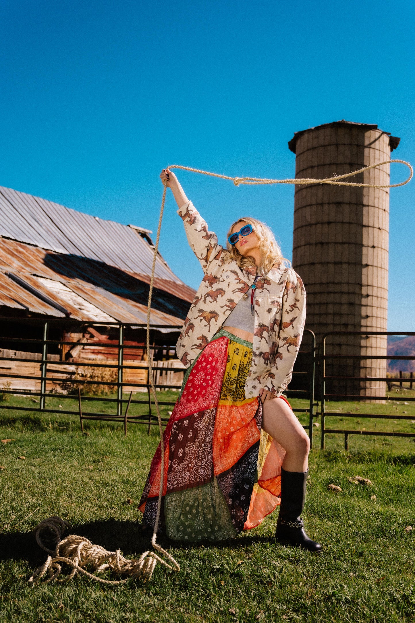 Person holding a lasso in front of a barn and silo on a sunny day wearing a patchwork style bandana print pleated midi skirt with a side slit