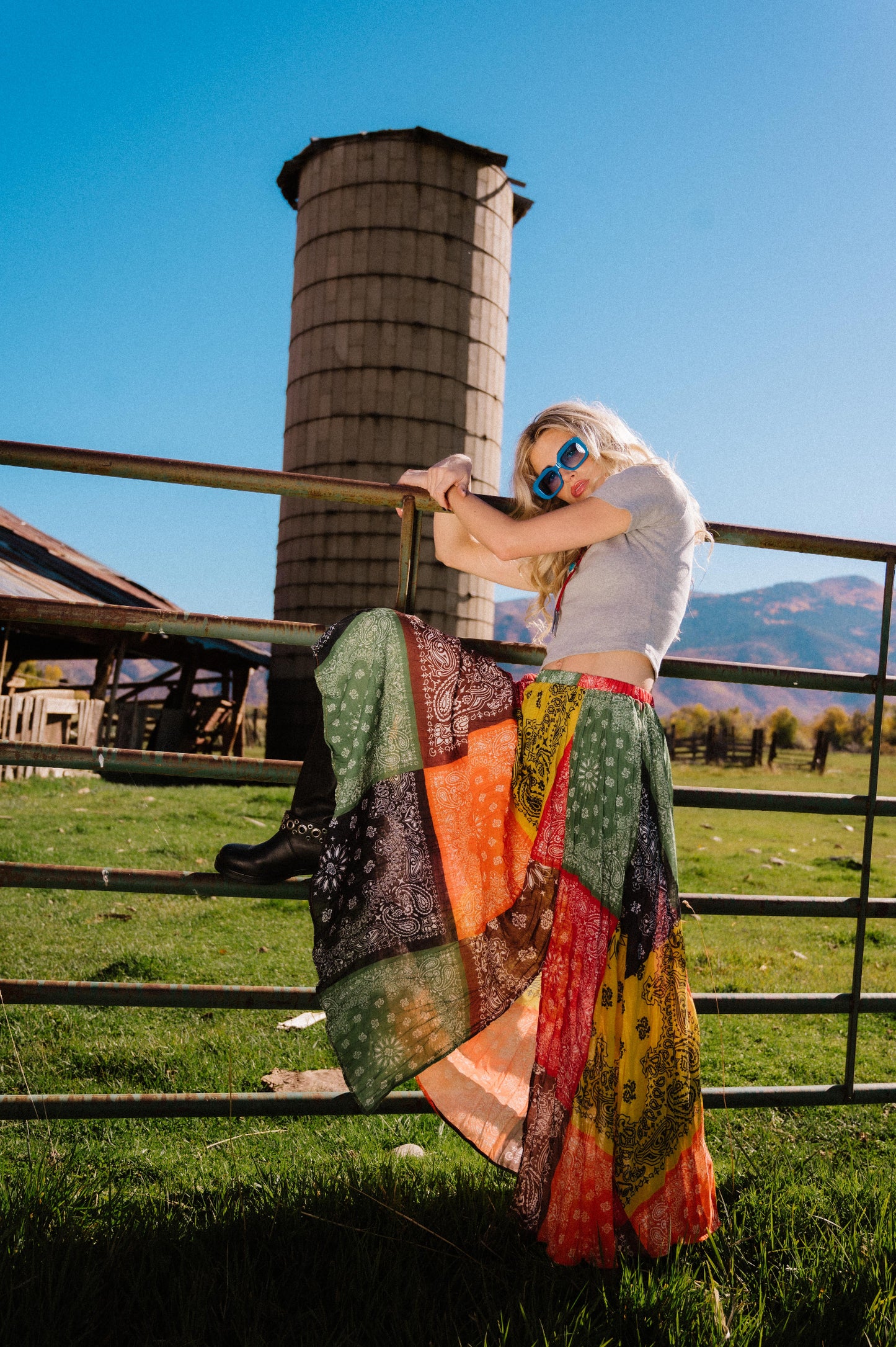 Person wearing a patchwork style bandana print pleated midi skirt with a side slit standing in front of a silo with mountains in the background