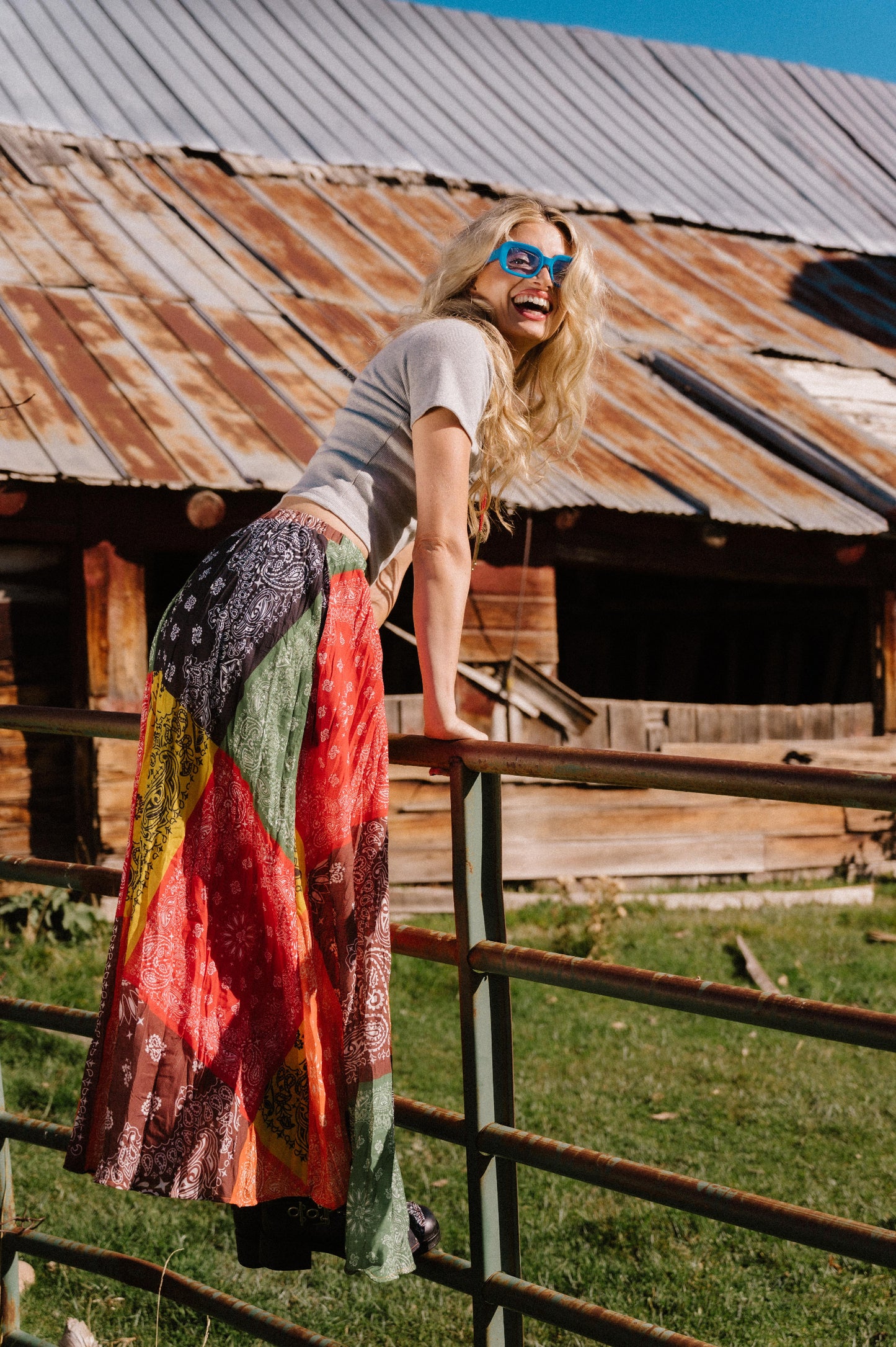 Woman standing on a wooden fence with a rustic building in the background wearing a patchwork style bandana print pleated midi skirt