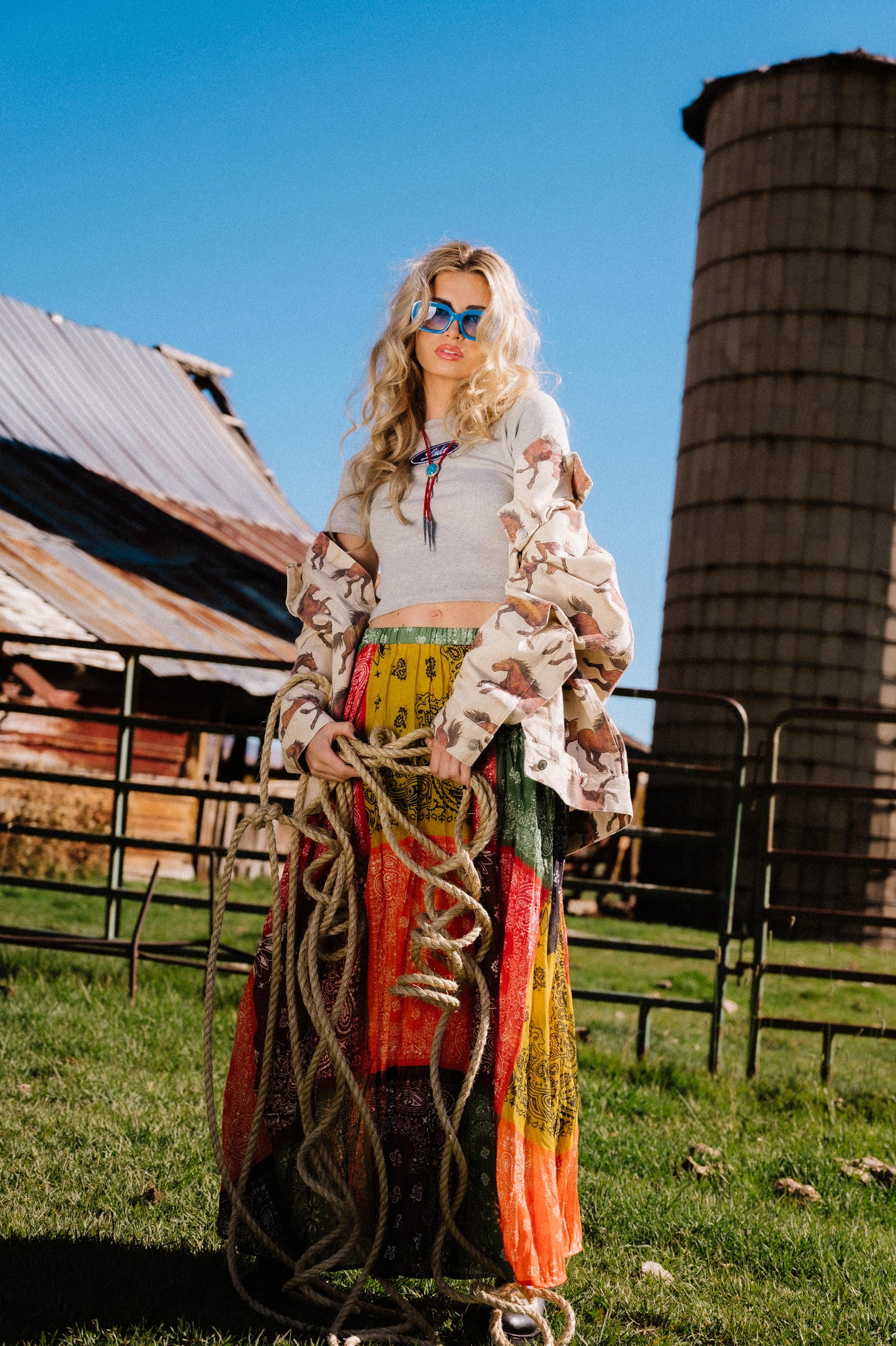 Woman holding a rope in front of a barn and silo wearing a patchwork style bandana print pleated midi skirt