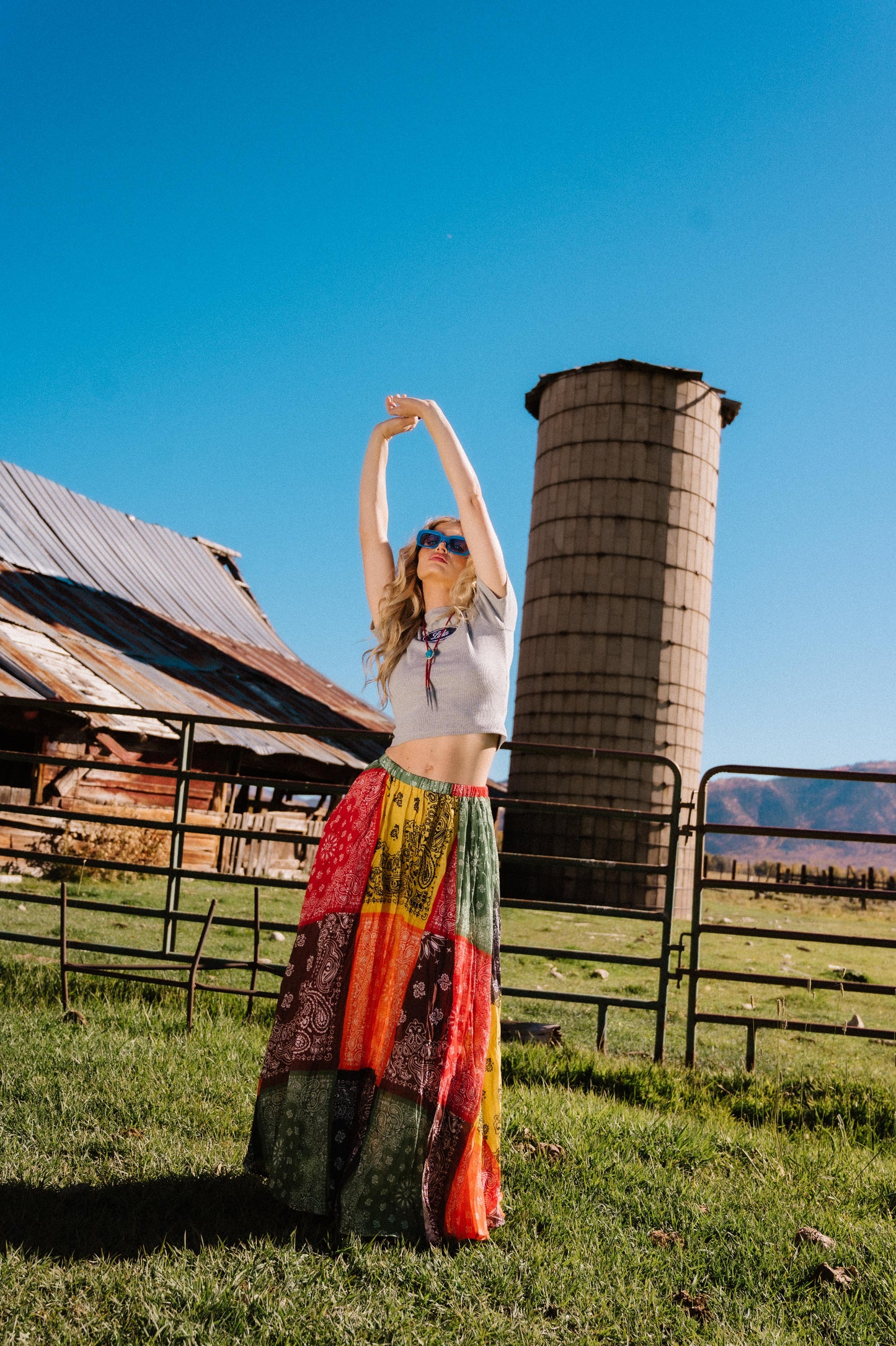 Person in patchwork style bandana print pleated midi skirt standing in a rural setting with a silo and barn in the background.
