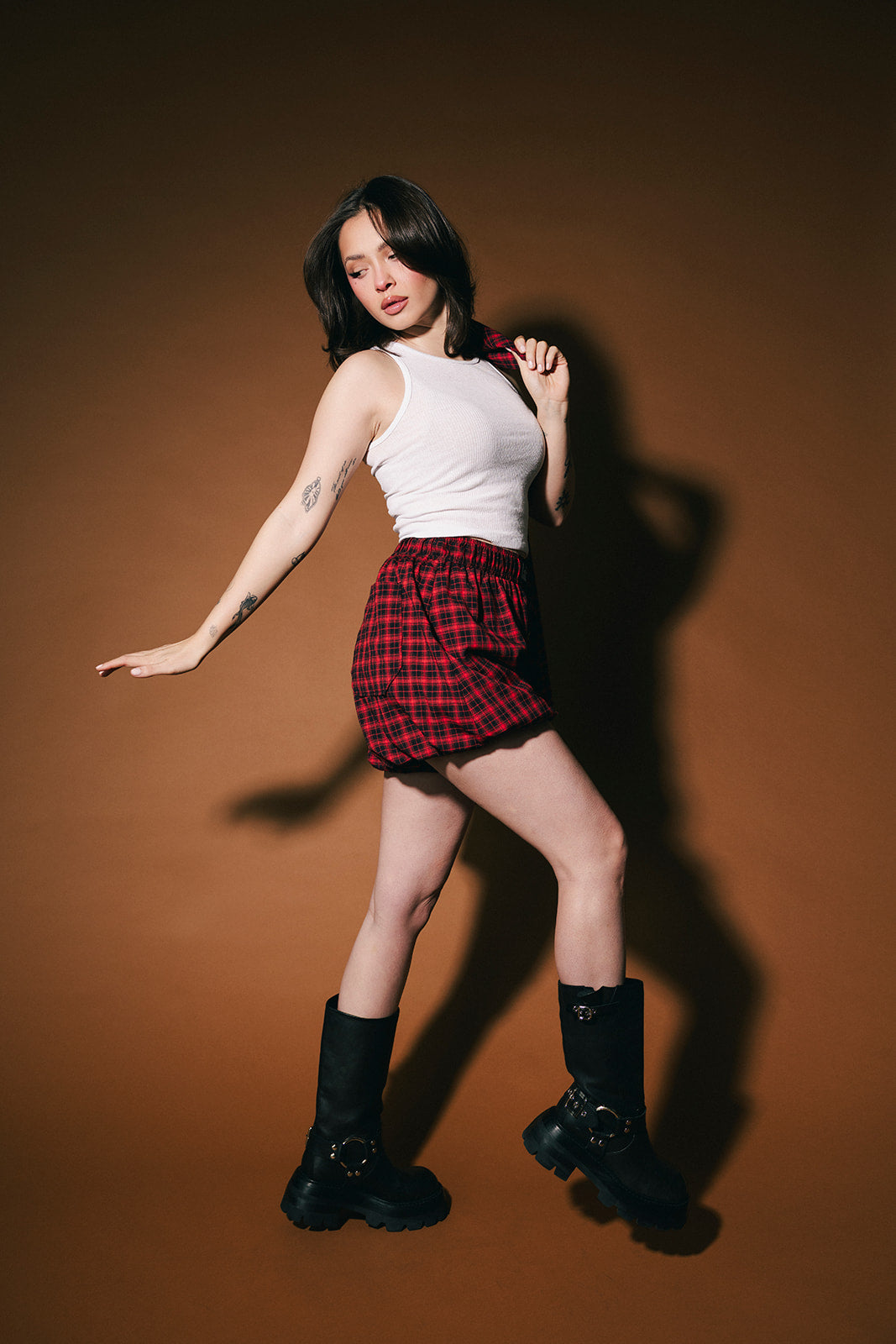 Woman posing in red plaid shorts and white top against a brown background