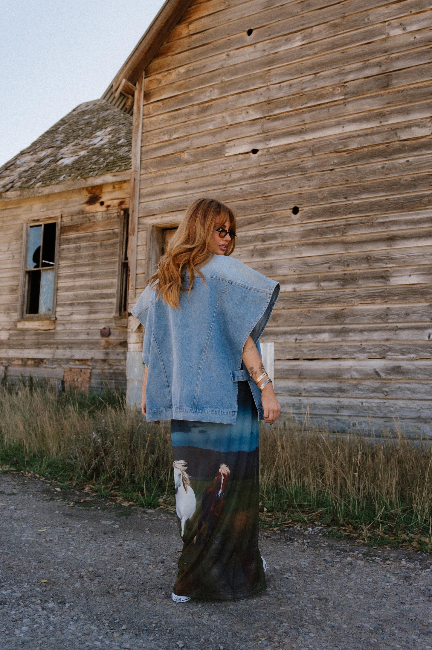 Woman standing in front of an old wooden building wearing a denim jacket and patterned dress.
