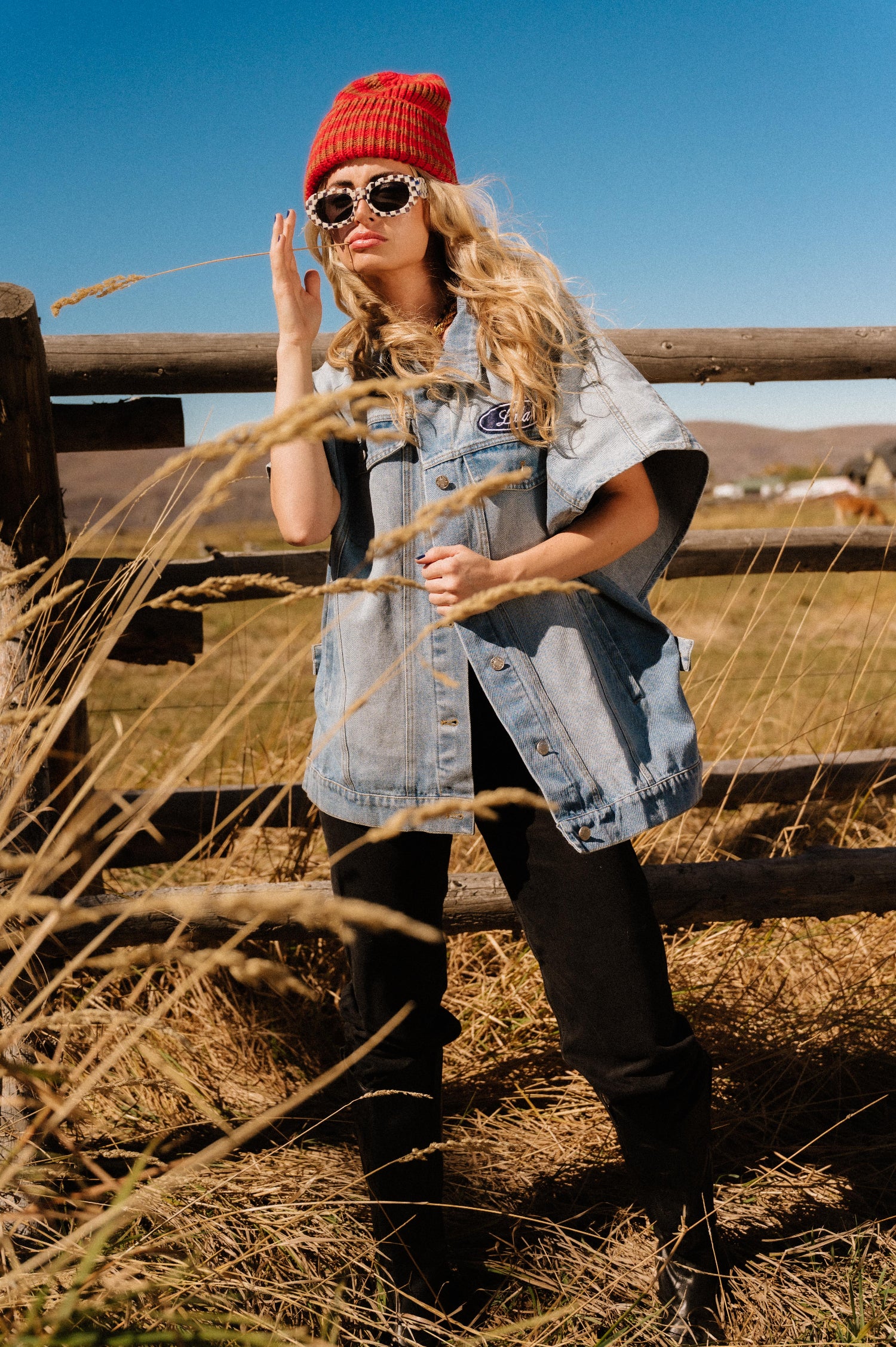 Woman in a red beanie and sunglasses standing in a field with a wooden fence and blue sky in the background.