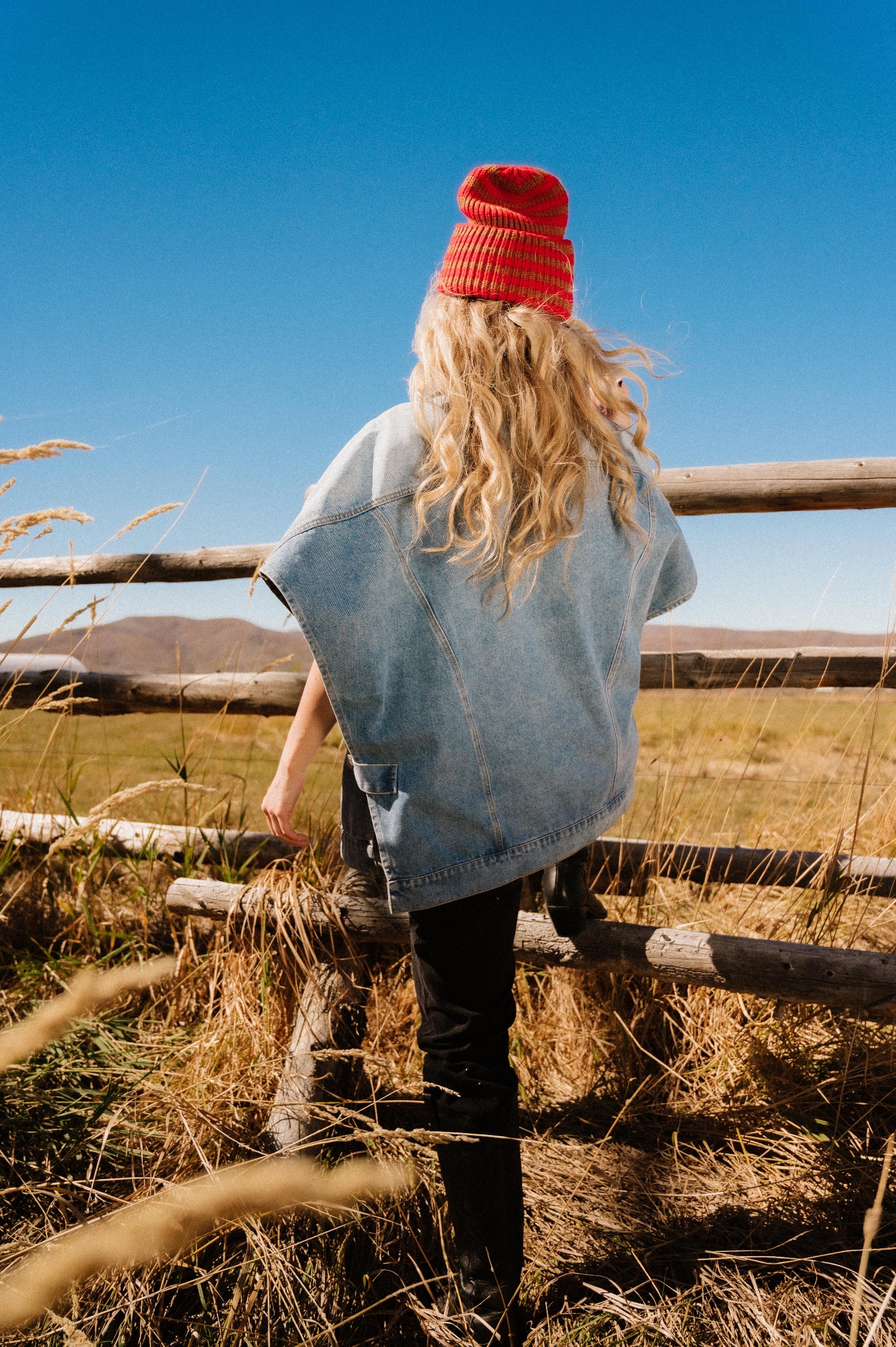Person wearing a red beanie and denim jacket standing on a wooden fence in a field with a clear blue sky.