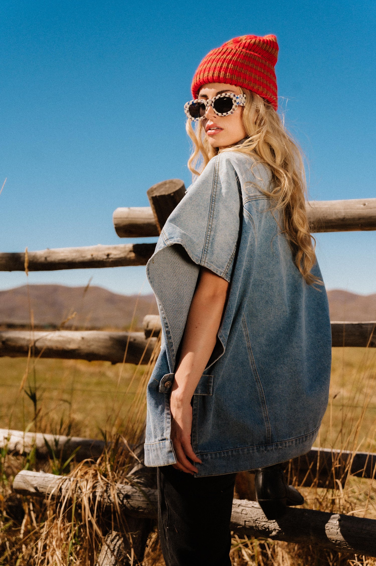 Woman wearing a red beanie and sunglasses, standing in a field with mountains in the background.
