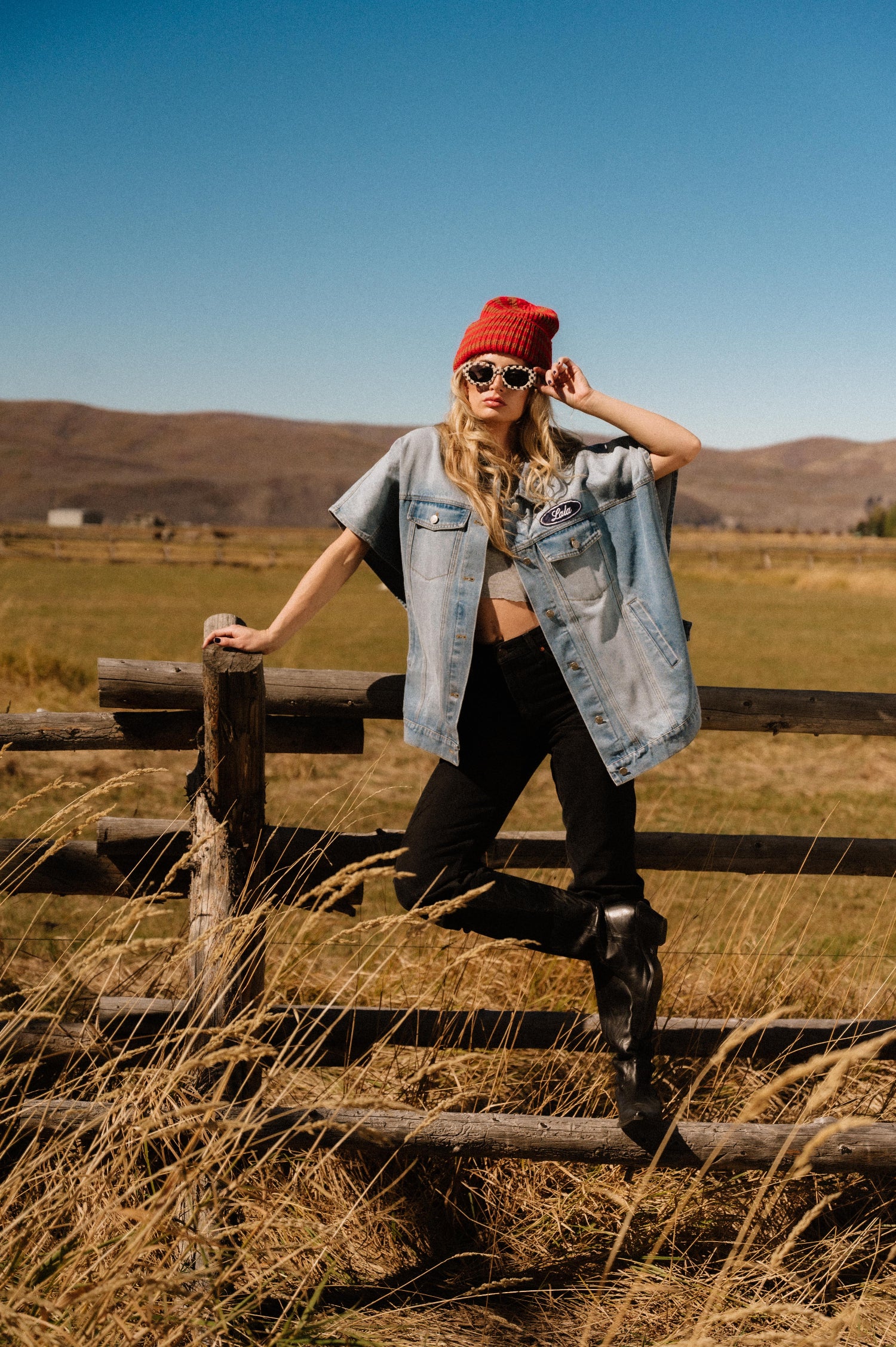 Person wearing a red beanie and sunglasses, standing on a wooden fence in a field with mountains in the background.