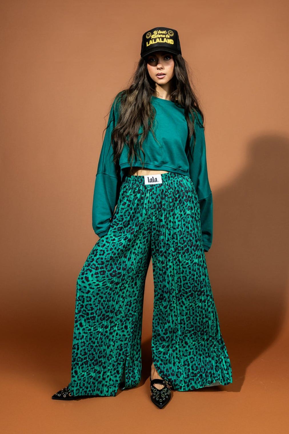 A girl/model standing in front of a backdrop wearing pleated green leopard print pants
