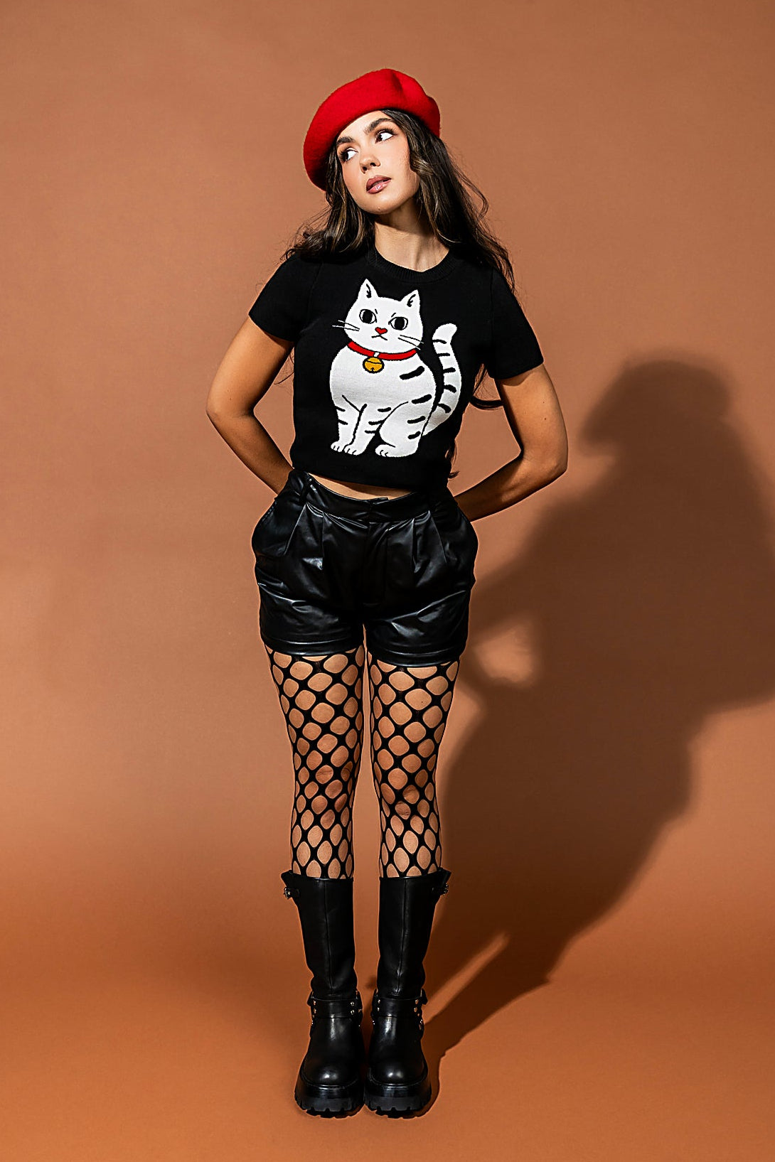 Woman wearing a black t-shirt with a cat graphic, black shorts, fishnet stockings, and a red beret on a brown background.