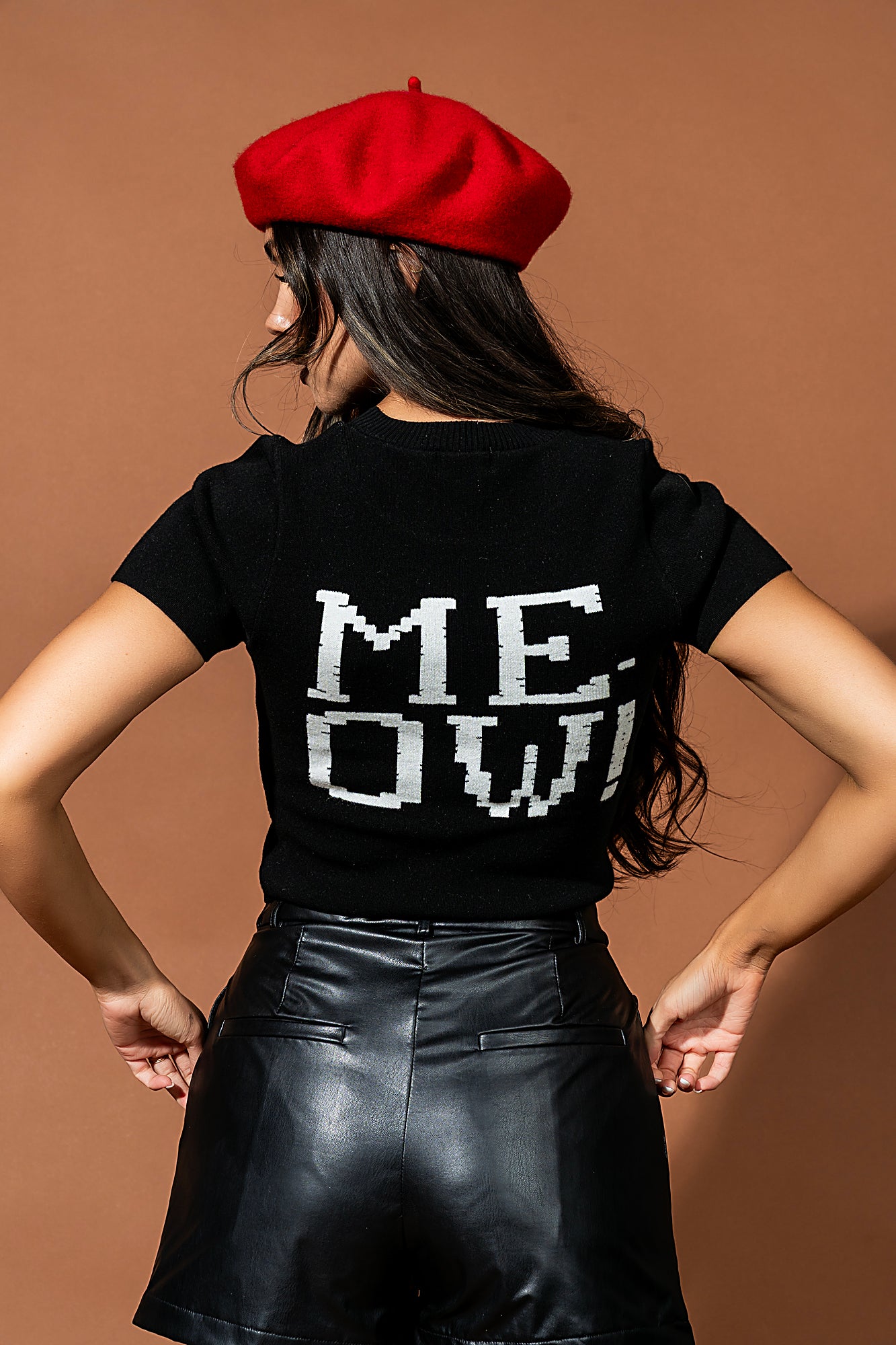 Person wearing a black t-shirt with 'MEOW' printed on it, red beret, and black leather skirt against a brown background