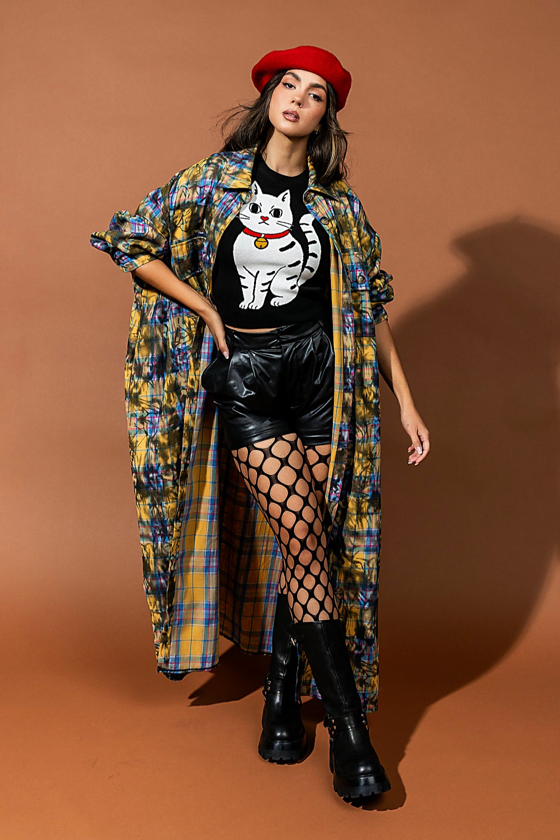Woman in stylish outfit with plaid shawl, black top with cat graphic, leather shorts, and black boots on a brown background