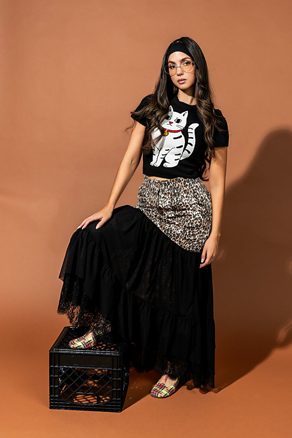 Woman wearing a black t-shirt with a white cat design, black skirt, and patterned top, sitting on a black crate against a brown background.