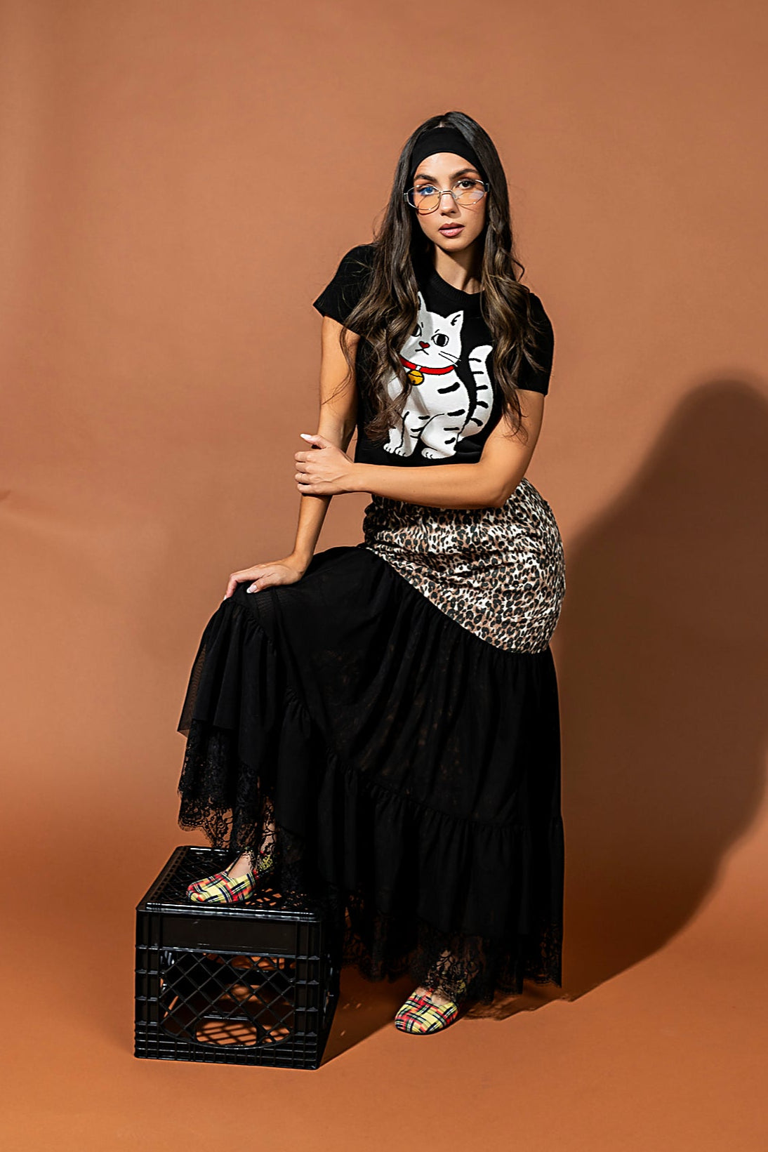Woman in black outfit with graphic t-shirt and skirt, sitting on a crate against a brown background