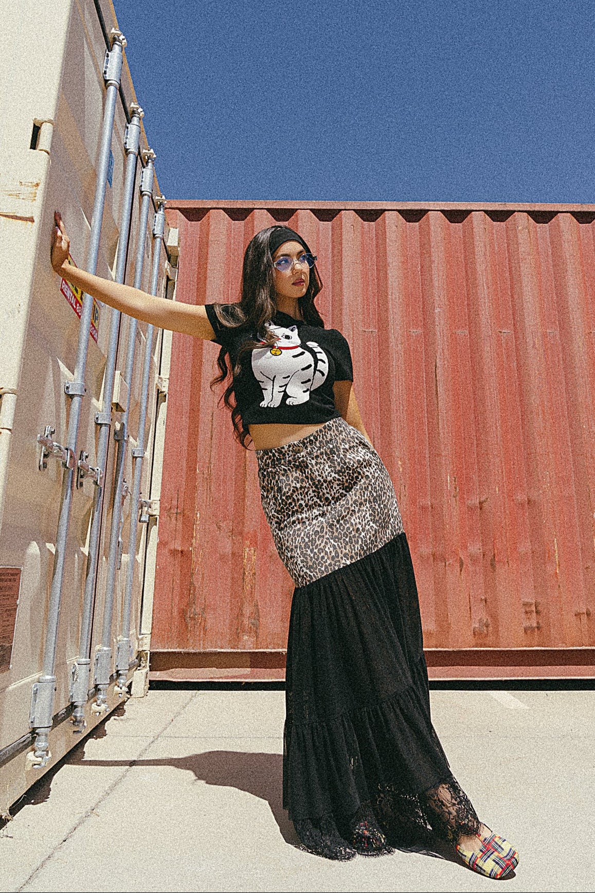 Woman standing in front of shipping containers wearing a black t-shirt with a graphic design and a long black skirt.