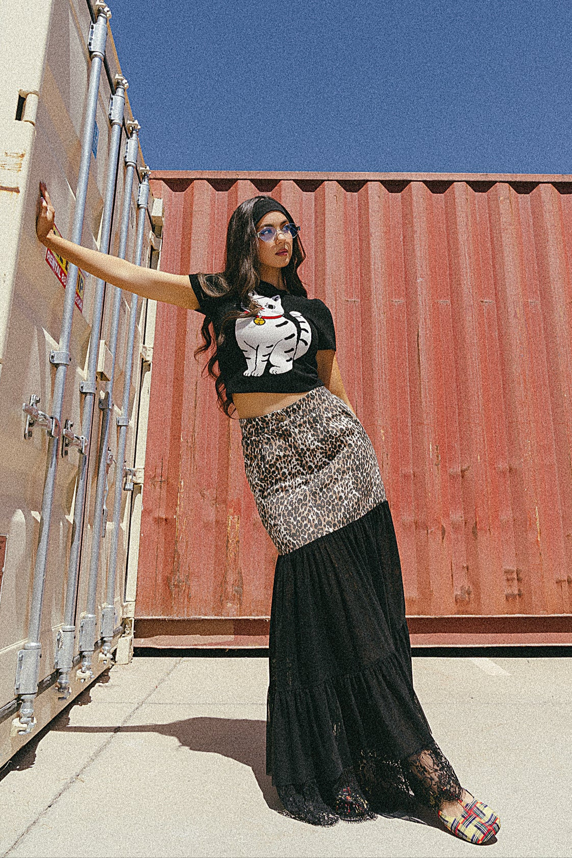 Woman standing in front of shipping containers wearing a black t-shirt with a graphic design and a long black skirt.