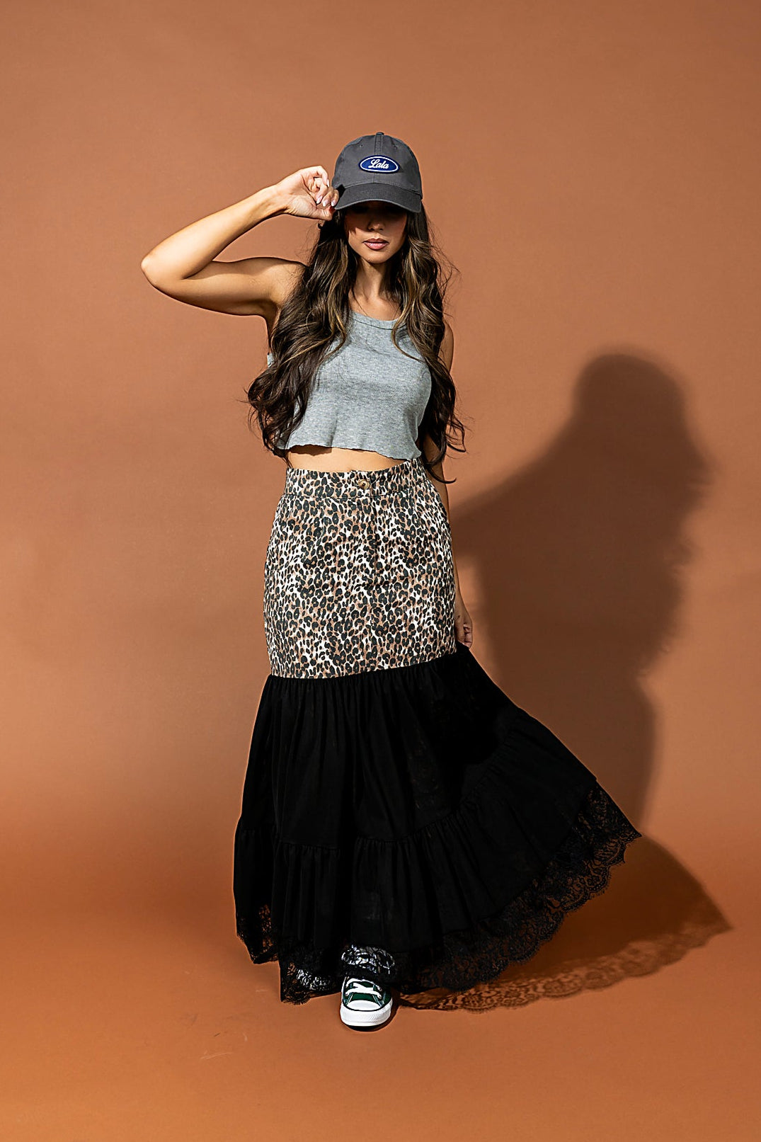 Woman posing in a gray crop top, black skirt, and blue cap against a brown background