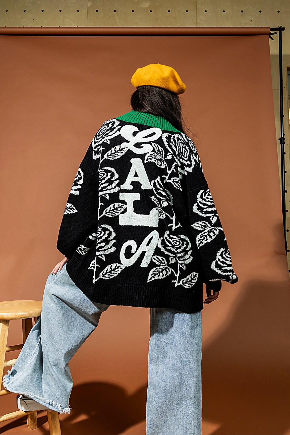Person wearing a black floral-patterned coat with white text, sitting on a stool against a brown backdrop.