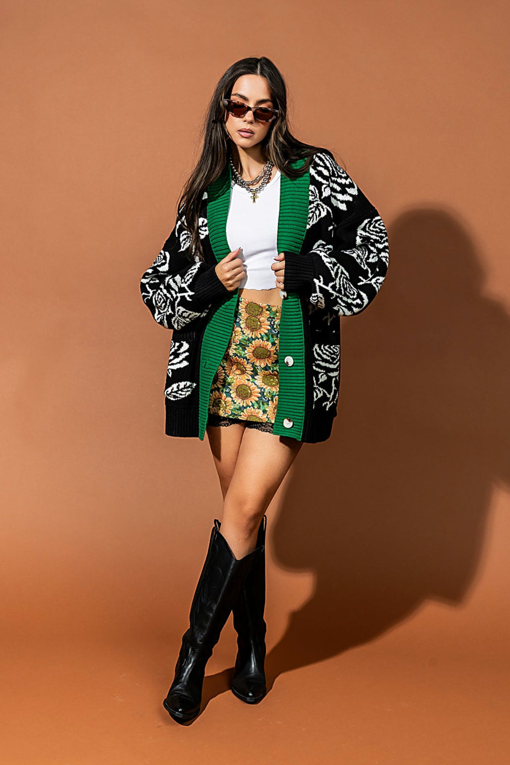 Woman wearing a green and black patterned cardigan, white top, floral skirt, and black knee-high boots against a brown background.