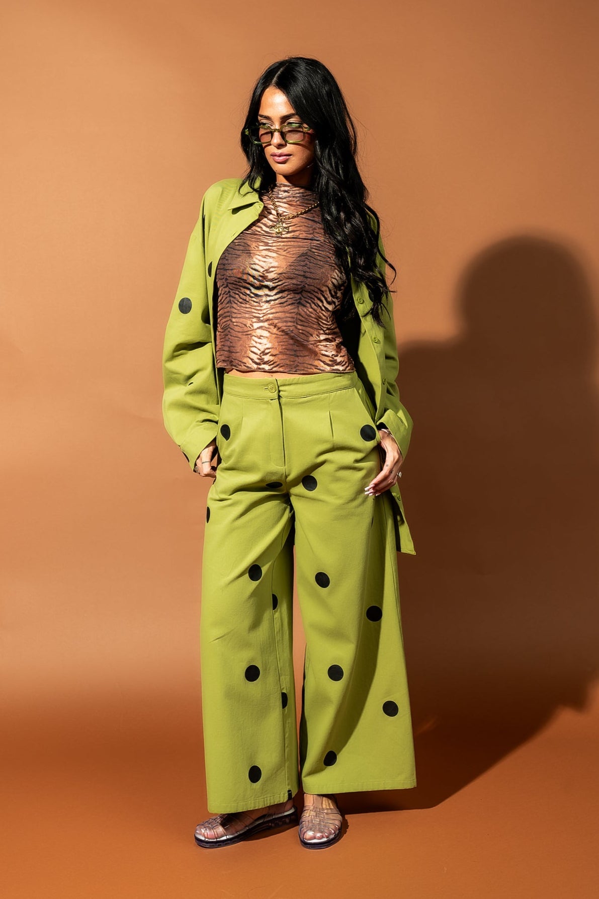 Girl/model standing against a brown backdrop wearing a tiger striped mesh top layered under a green shirt jacket with black polka dots