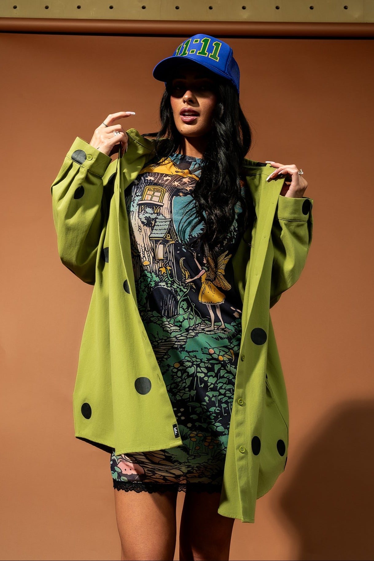 Girl/model standing against a brown backdrop wearing a green shirt jacket with black polka dots, a mesh dress with a fairy pattern on it and a blue trucker hat with 11:11 graphics