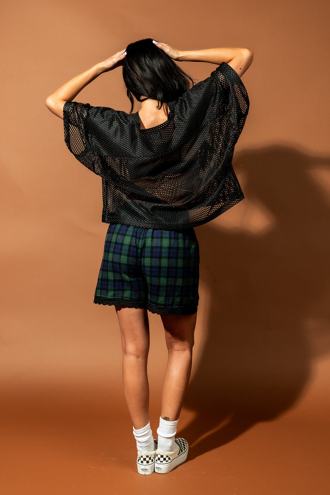 Girl/model wearing a black football jersey with a big yellow smile face printed on it with blue and green plaid shorts, facing away