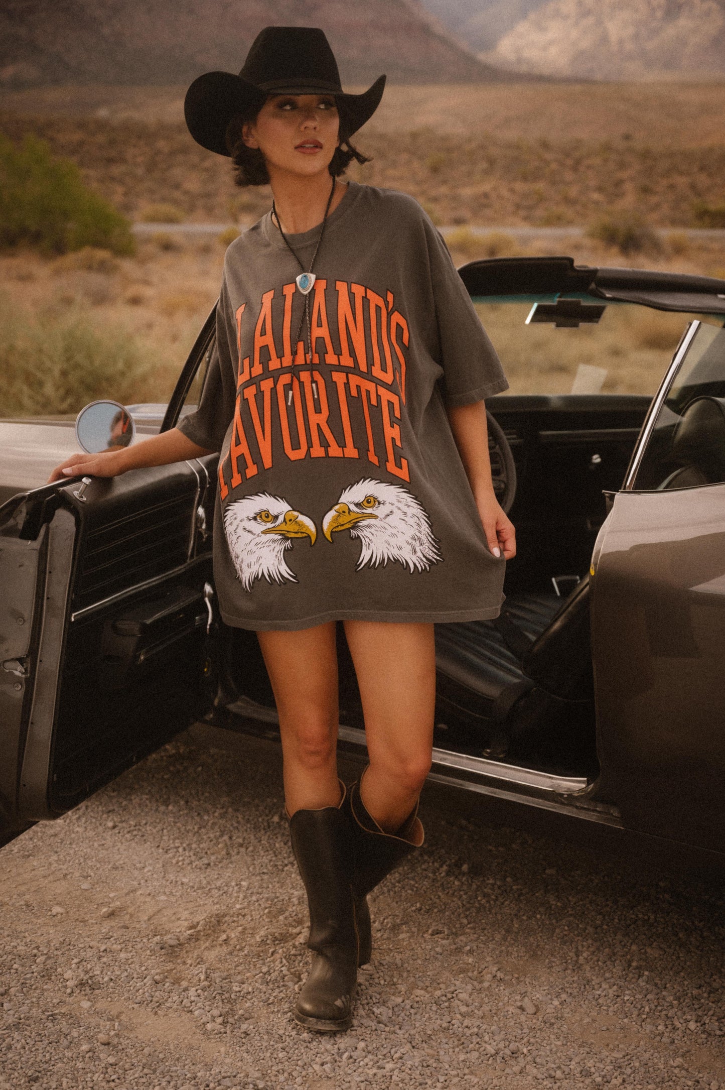 Person wearing a graphic t-shirt and cowboy hat standing next to an open car in a desert setting