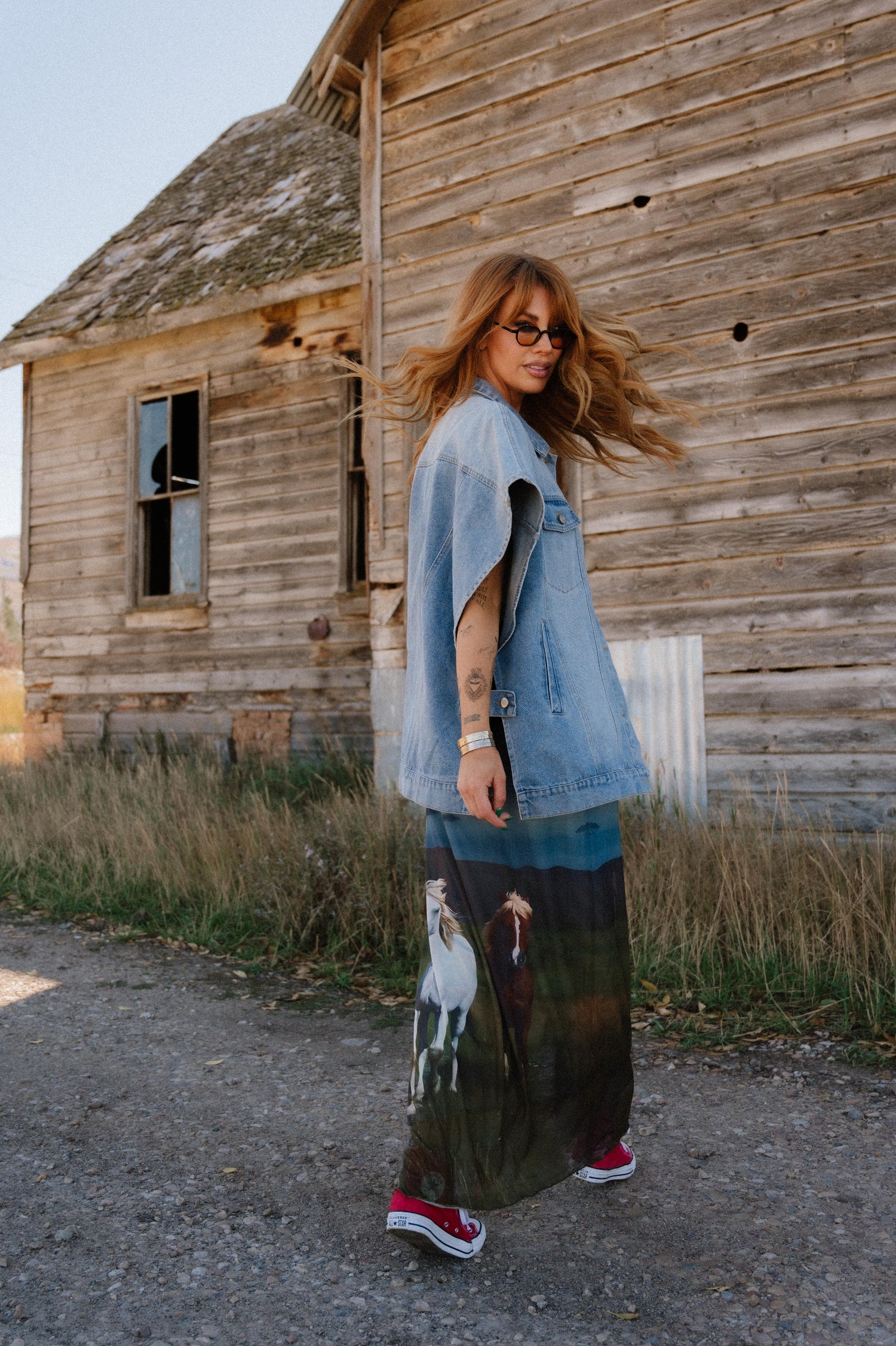Woman standing in front of an old wooden building wearing a denim jacket and patterned dress.