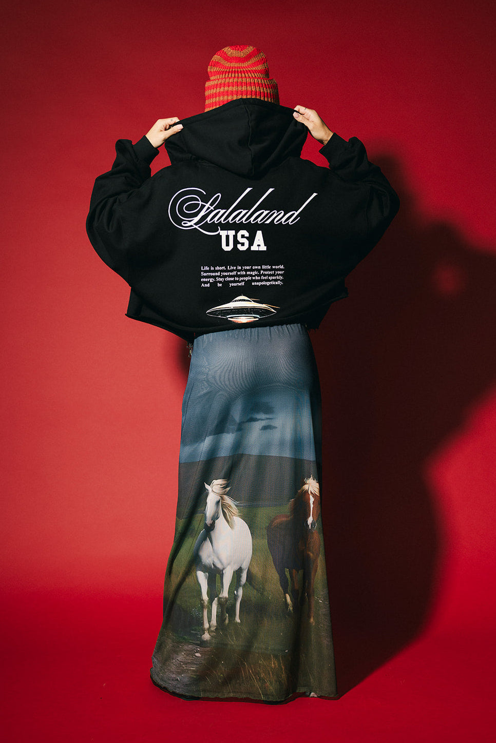 Person wearing a black hoodie with 'LALALAND USA' branding and a long skirt with horse print against a red background.