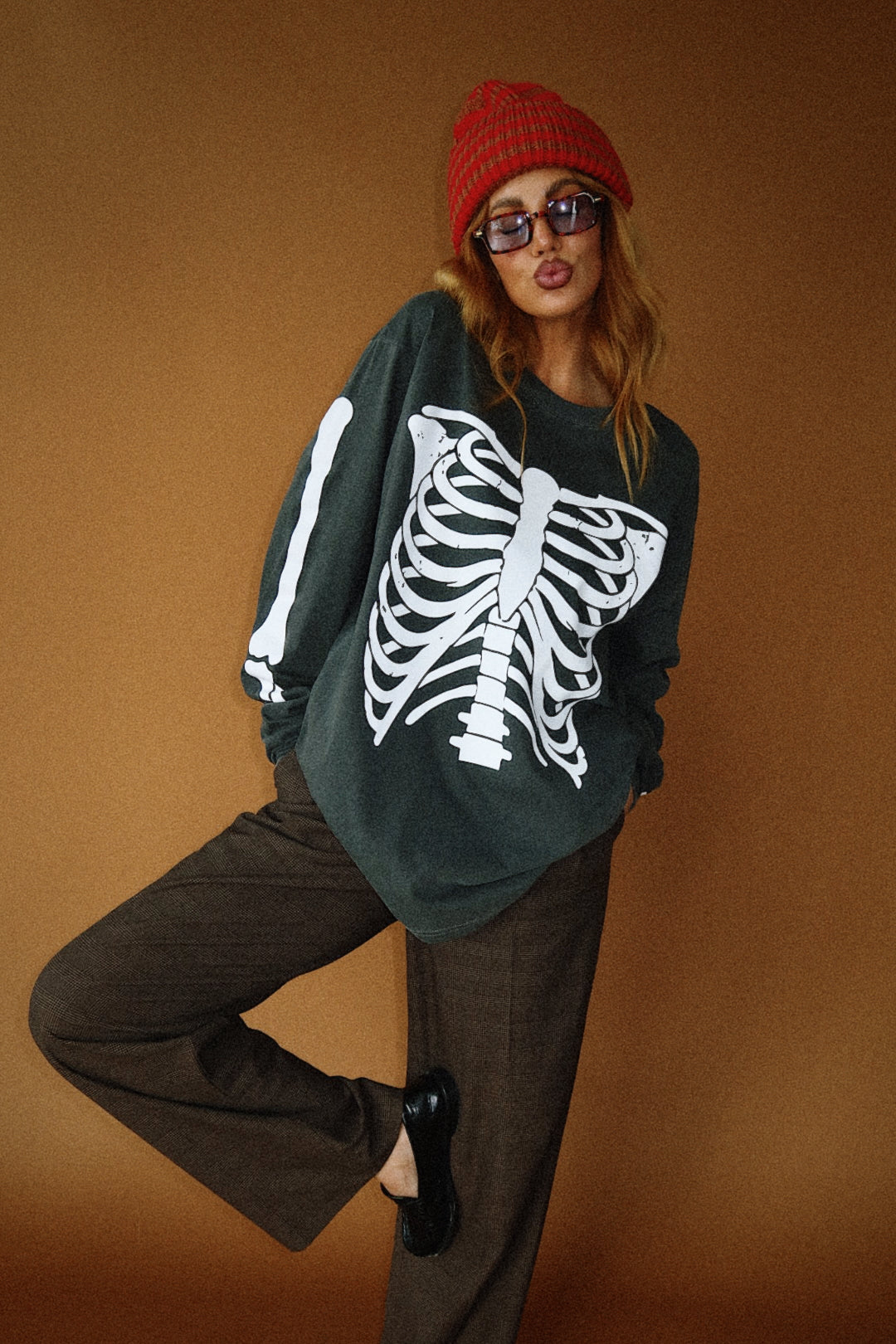 Person wearing a dark grey sweater with a white skeleton design, brown pants, and a red beanie on a brown background.