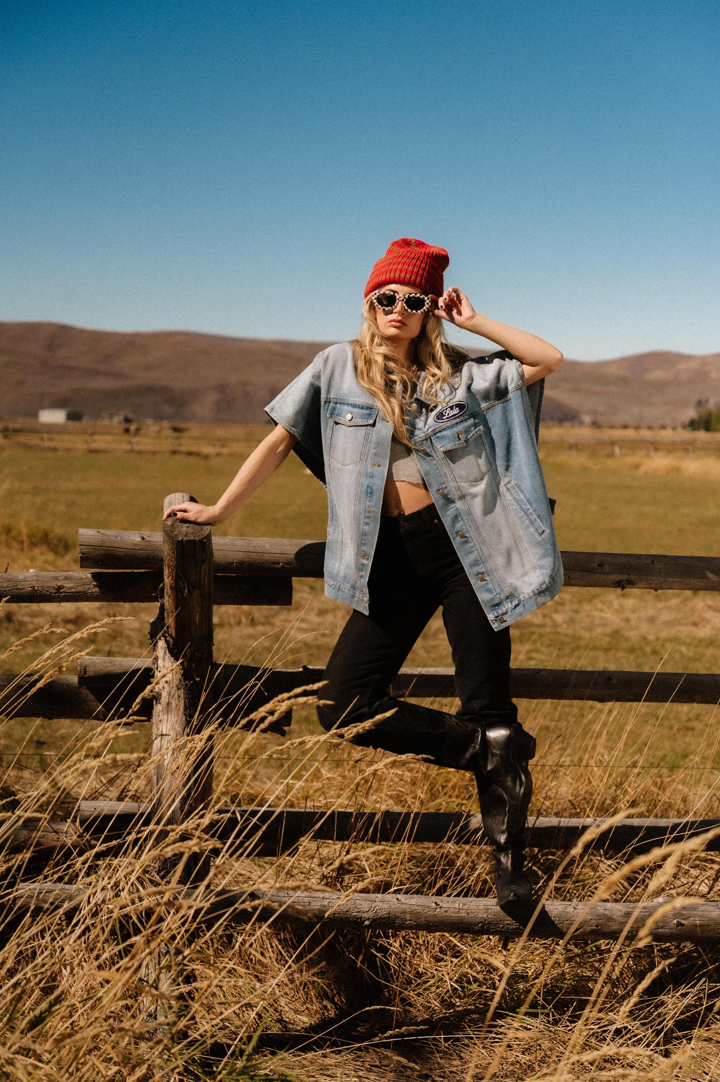 Person wearing a red beanie and sunglasses, standing on a wooden fence in a field with mountains in the background.