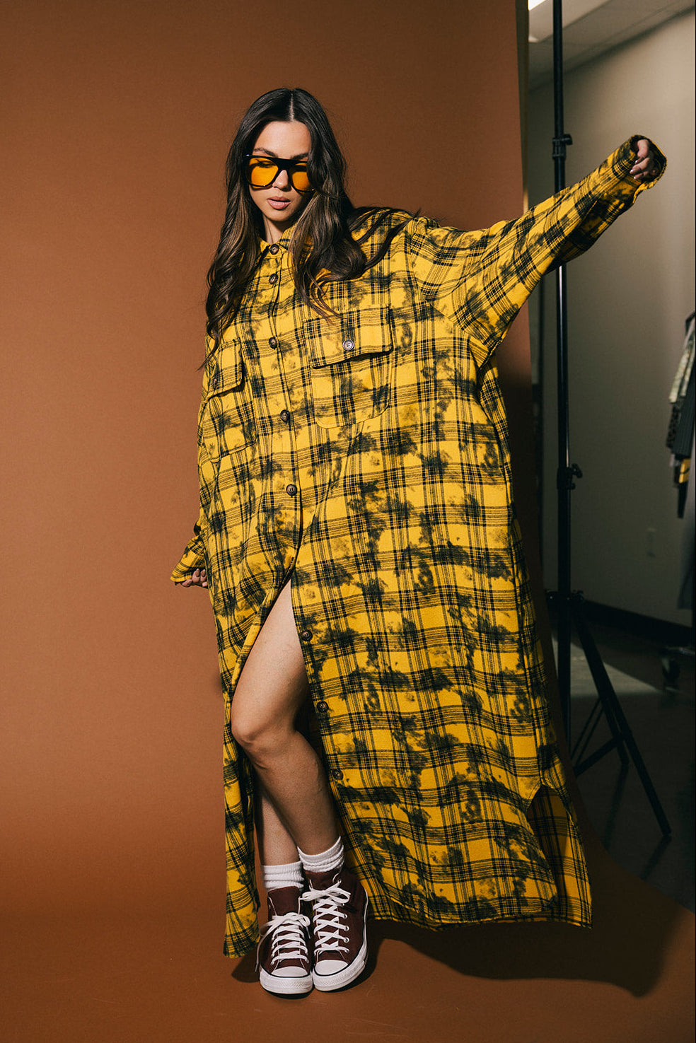 Woman wearing a yellow plaid dress, standing against a brown background.