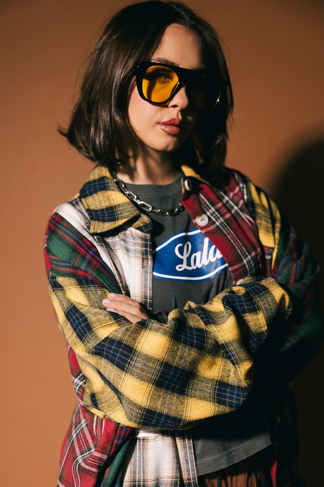 Person wearing a long patchwork plaid duster over a graphic t-shirt with sunglasses on a brown background
