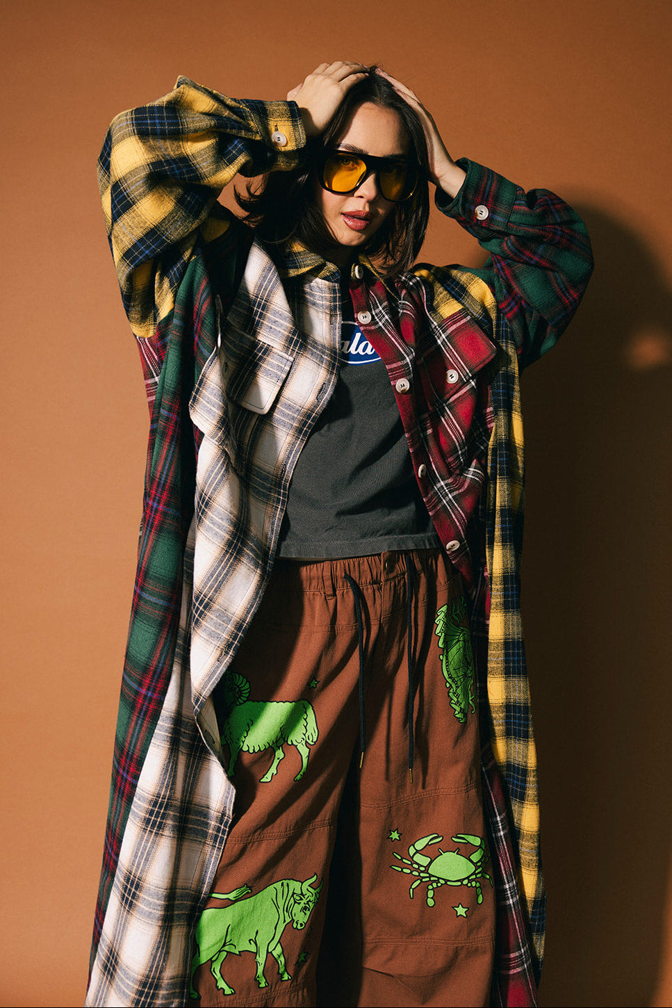 Person wearing a patchwork plaid duster over a graphic t-shirt with brown pants featuring green designs, against a brown background.