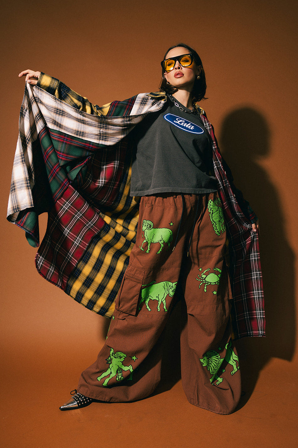 Person wearing a patchwork plaid duster over a graphic t-shirt with brown pants featuring green animal prints on a brown background