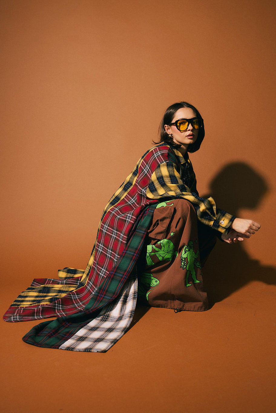 Person wearing a patchwork plaid duster and sunglasses on a brown background