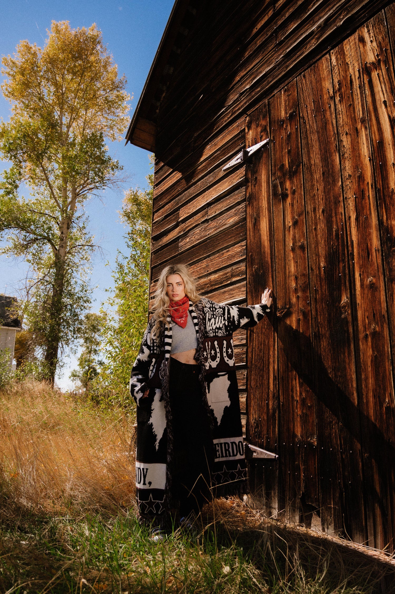 Person wearing a patterned cardigan standing next to a wooden building with trees in the background