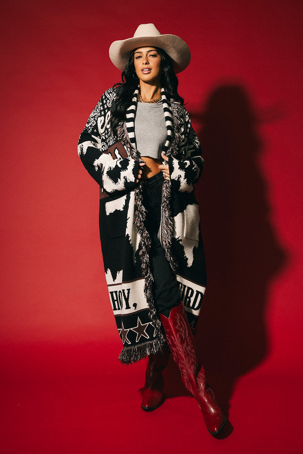 Woman wearing a patterned cardigan, white hat, and red boots against a red background