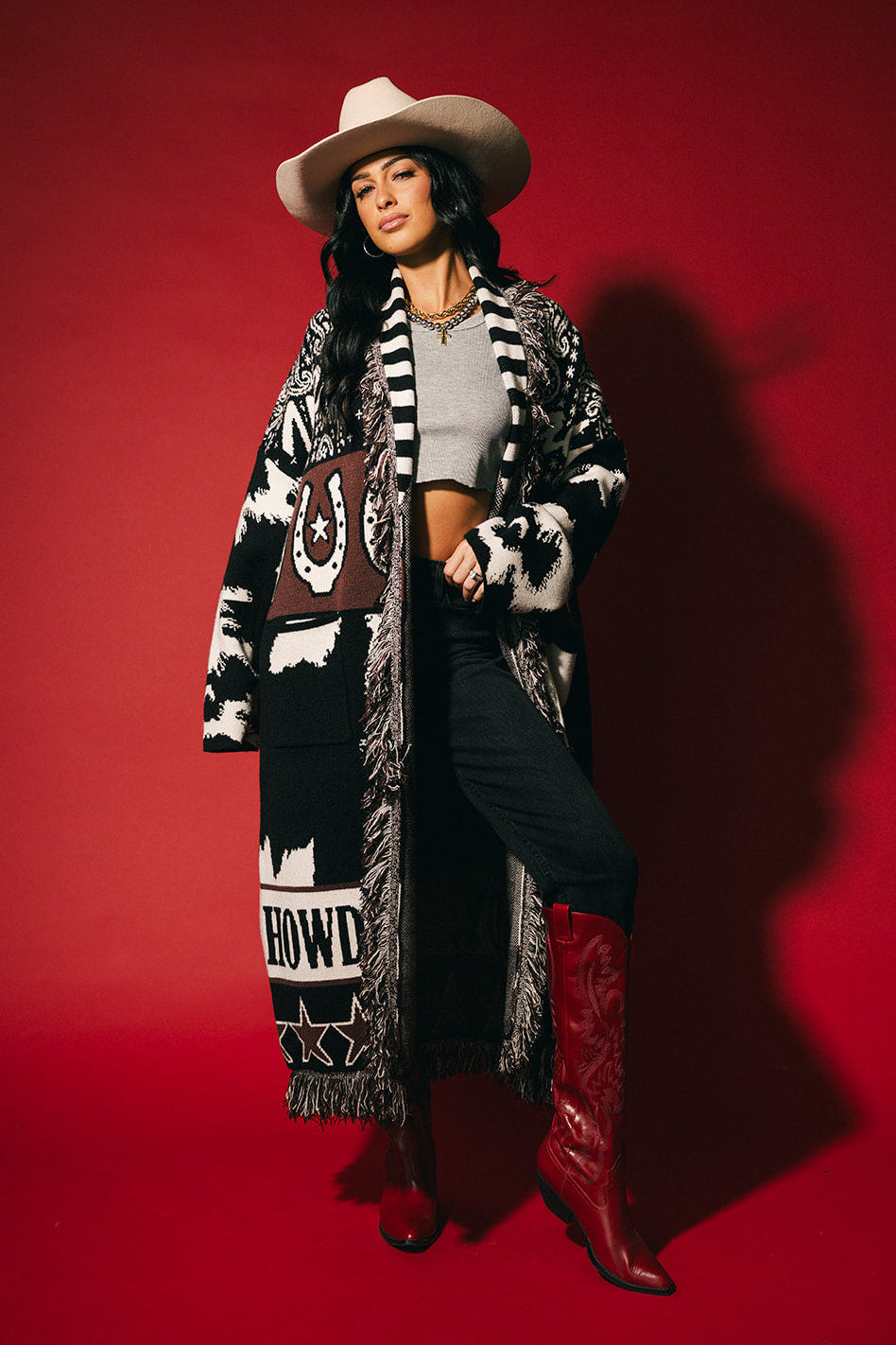 Person wearing a long black and white patterned cardigan, red boots, and a cowboy hat on a red background