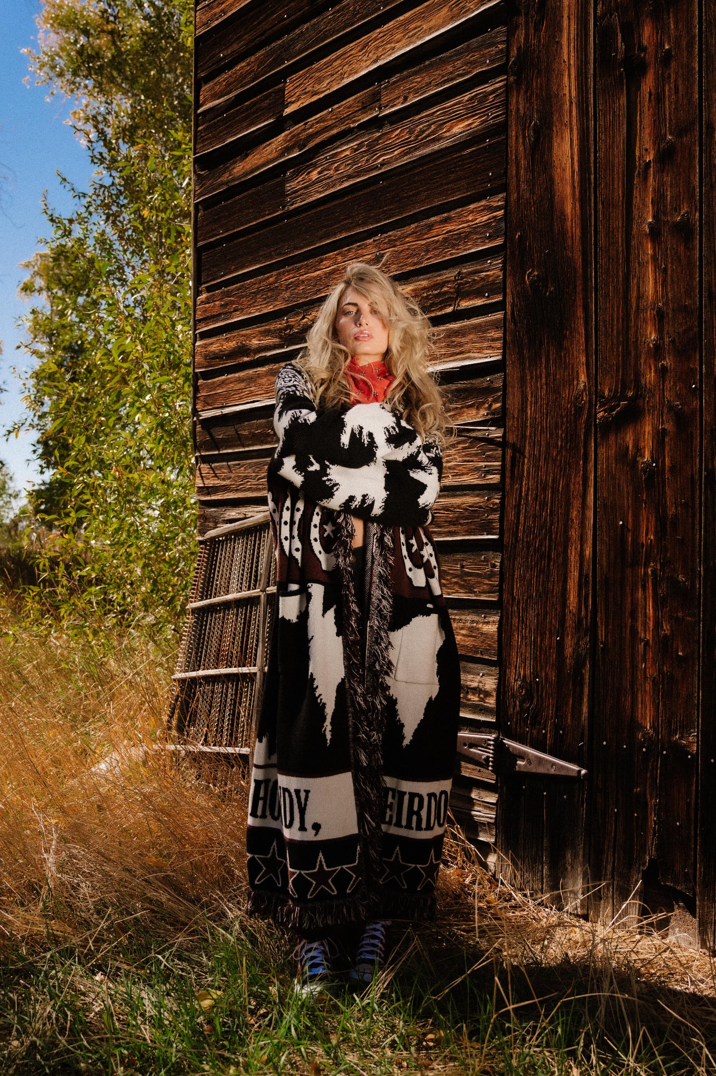 Person wearing a long, patterned cardigan standing in front of a wooden building with trees in the background.