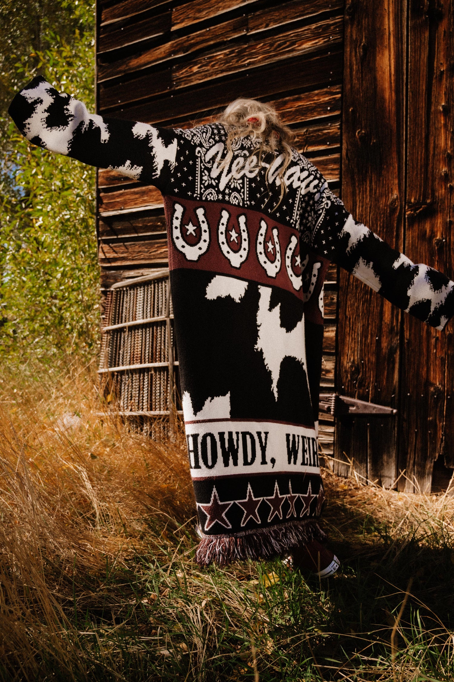 Person wearing a patterned sweater with text and graphics in front of a wooden building.