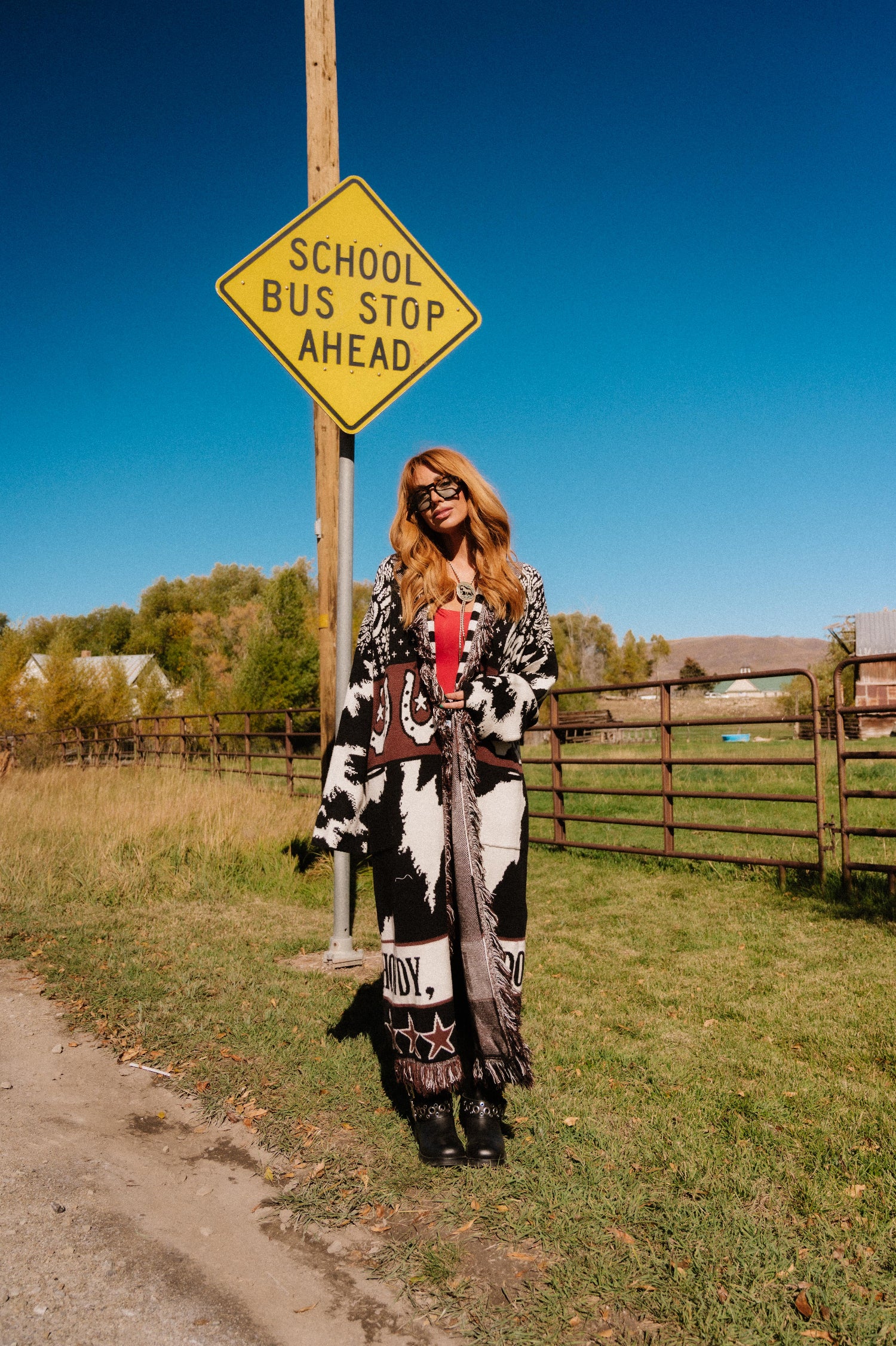 Person in a patterned outfit standing next to a 'School Bus Stop Ahead' sign in a rural setting.