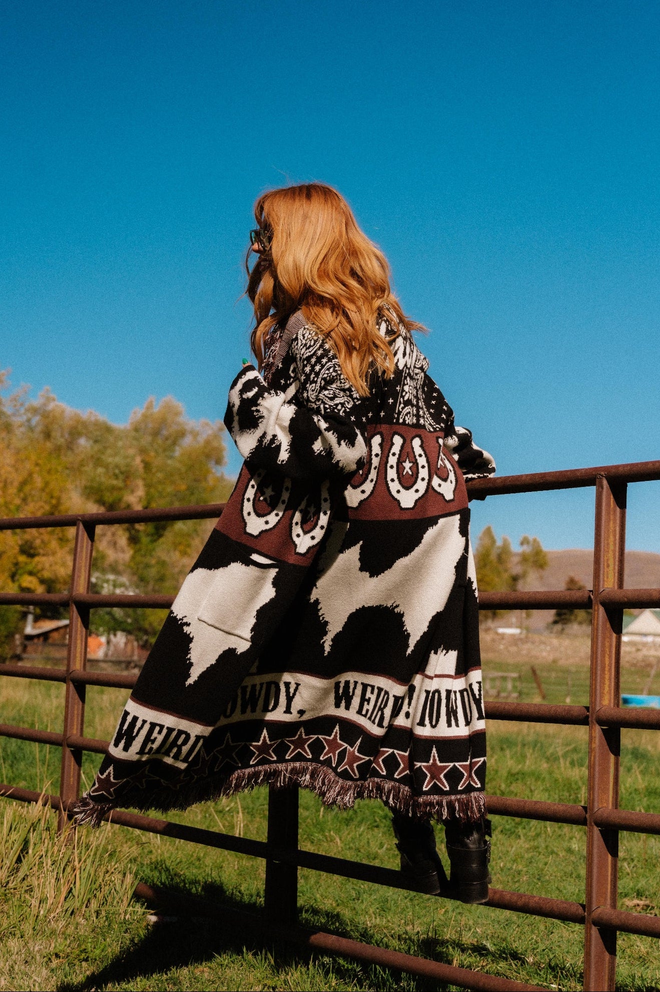 Person wearing a cowhide patterned cardigan standing by a fence with a clear blue sky and trees in the background.