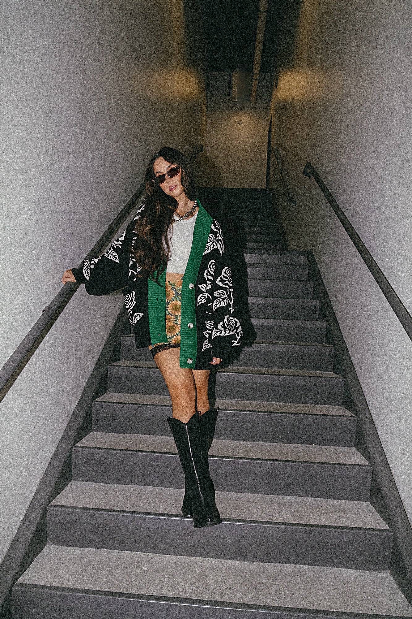Woman walking down a set of stairs wearing a black and white floral cardigan, green scarf, and black knee-high boots.